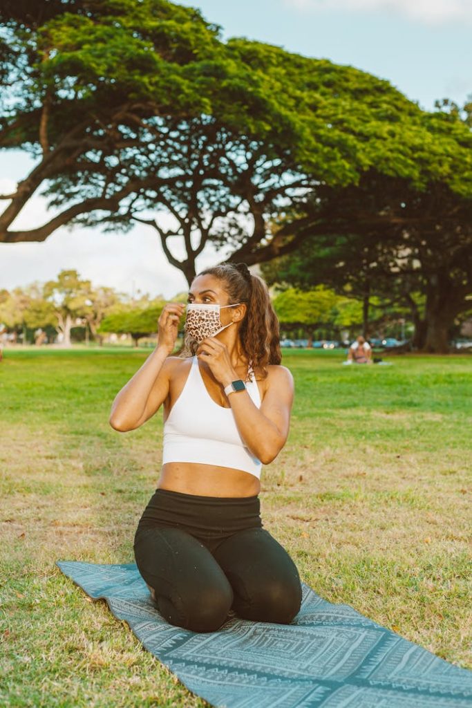 Woman kneeling on yoga mat in park, adjusting face mask, blending fitness with safety.